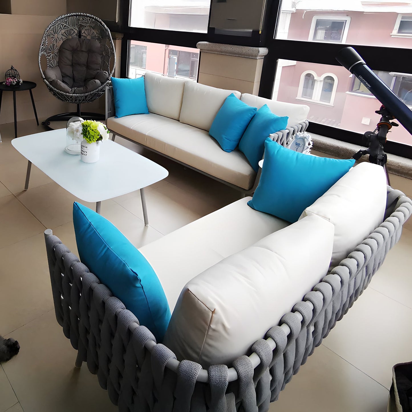 The Elysian Breeze Collection by Landmark Decor features two gray woven couches with weather-resistant cushions and blue accent pillows. A white coffee table with a centerpiece rests between them, a telescope is by the window, a unique chair sits in the corner, and large windows provide natural light.