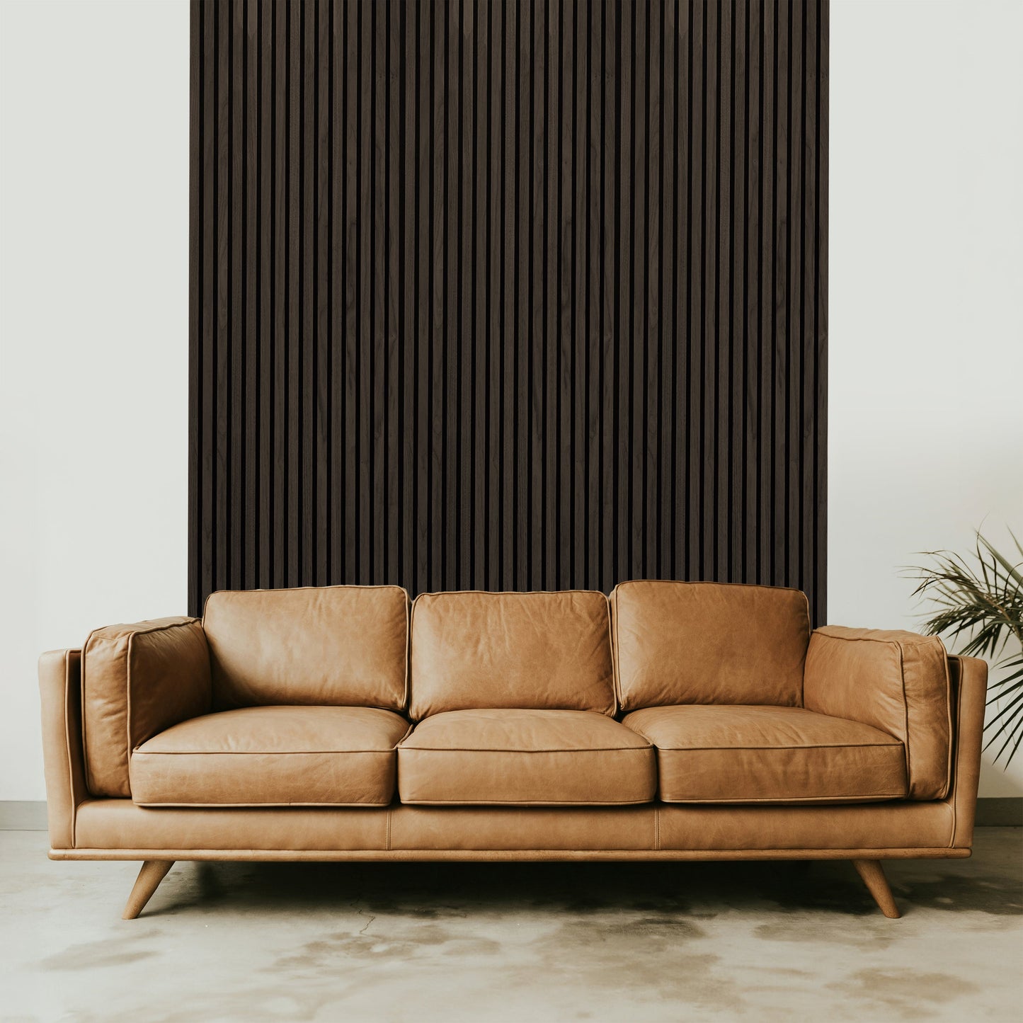 A brown leather sofa sits before a Landmark Decor Dark Smoke Oak Acoustic Slat Wall Panel, while a green plant on the right adds a refined touch to the interior design.