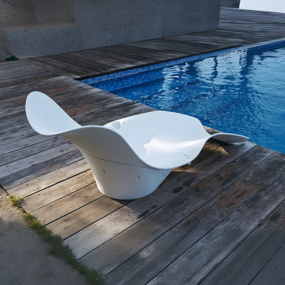 The Muse Astratta Lounger by Landmark Decor graces the wooden deck beside a blue pool, creating a luxurious retreat. Its modern sculptural design, with sleek white curves, embodies outdoor living, offering a striking contrast to the natural wood and shimmering water.