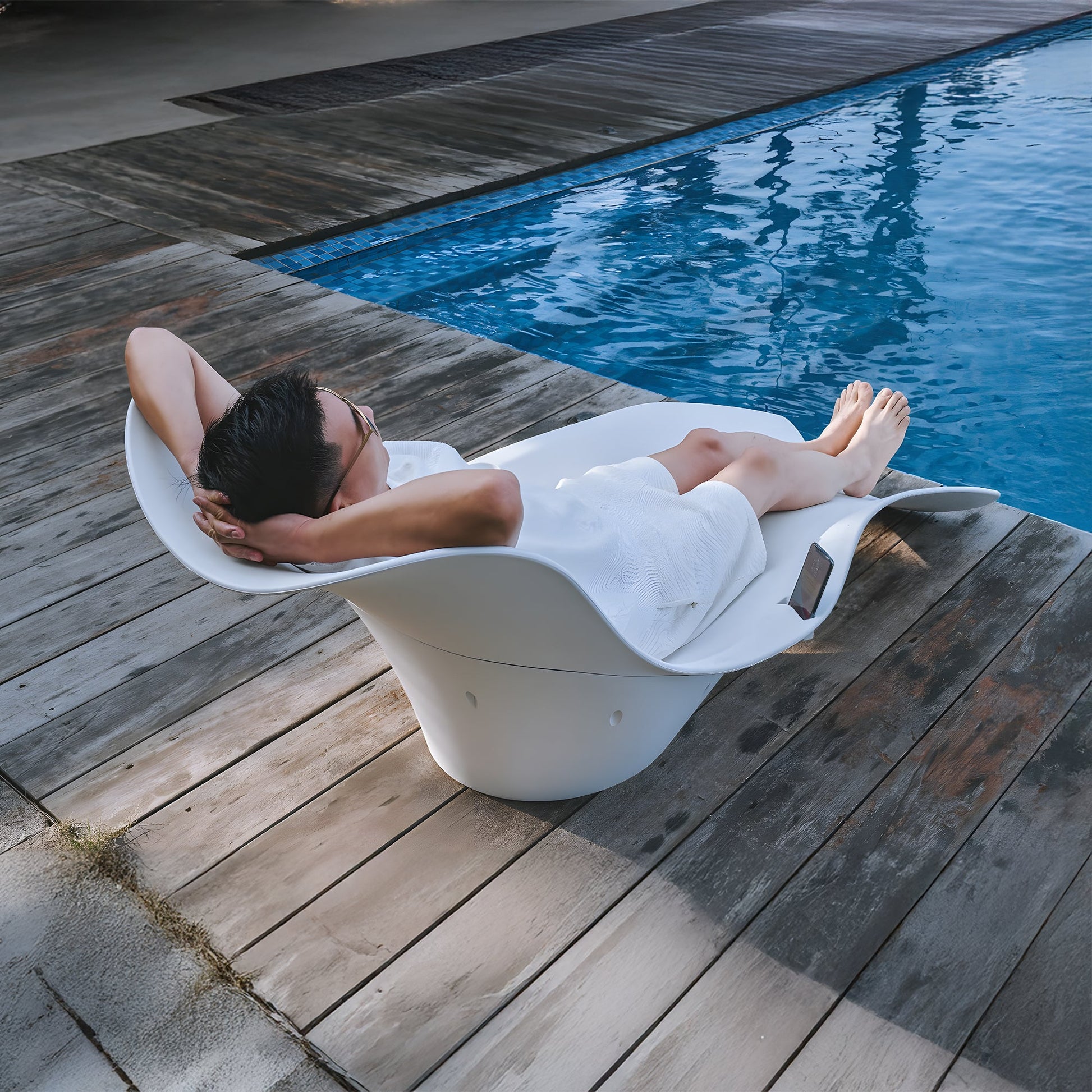Unwind in style on the Muse Astratta Lounger by Landmark Decor, lounging by a serene pool. With sunglasses on and hands behind your head, savor outdoor living while your smartphone sits nearby on the wooden deck.