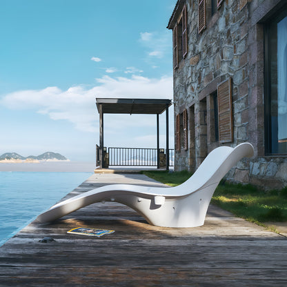 The Muse Astratta Lounger by Landmark Decor sits elegantly on a wooden deck by calm water, next to a stone house with wooden shutters. A small canopy provides shade, adding luxury to the retreat, while mountains rise in the distance under a clear blue sky.