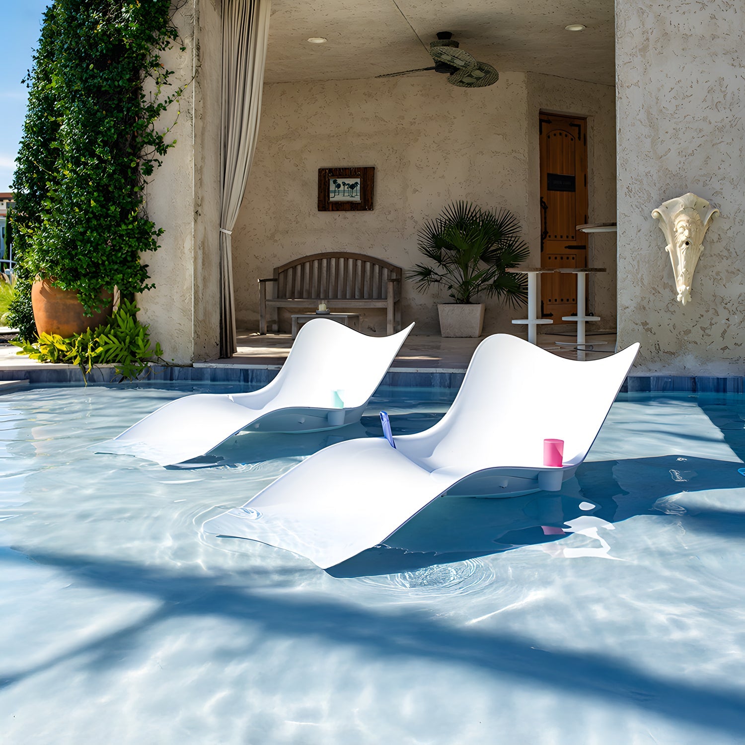 Two Muse Astratta Loungers by Landmark Decor partially submerged in a pool each hold a colorful cup, epitomizing outdoor living. Behind them is a shaded patio with a wooden bench, plants, fan, and decorative wall art. Sunlight casts inviting shadows across this luxurious retreat.