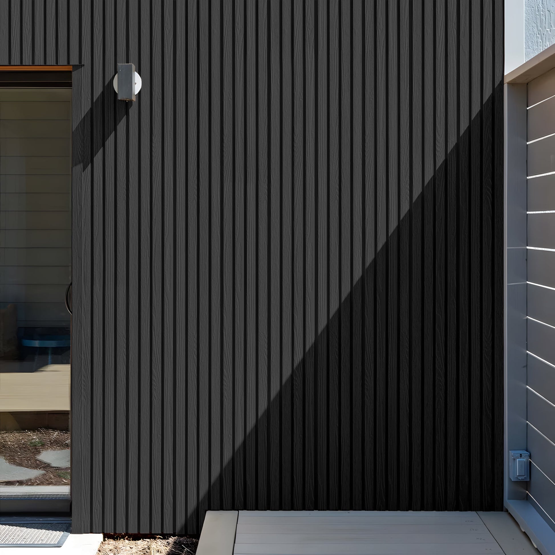 A minimalist exterior wall features Landmark Decor’s Urban Shadow Exterior WPC Panel in weather-resistant vertical dark panels, a compact light fixture, a glass door on the left, and sunlight casting a sharp diagonal shadow.