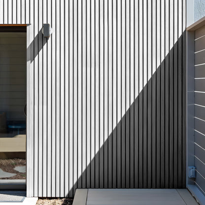 A modern facade features Arctic Frost Exterior WPC Panels by Landmark Decor in vertical slats, a wall-mounted light, a glass door on the left, and a sharp shadow dividing the wall diagonally.