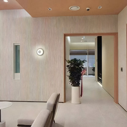 A modern minimalist interior features beige walls, a round light fixture, a tall potted plant, and a doorway. Lightweight Oman Linear Stone Sheet - Grey by Landmark Decor accents the space, with two light chairs partially visible in front.