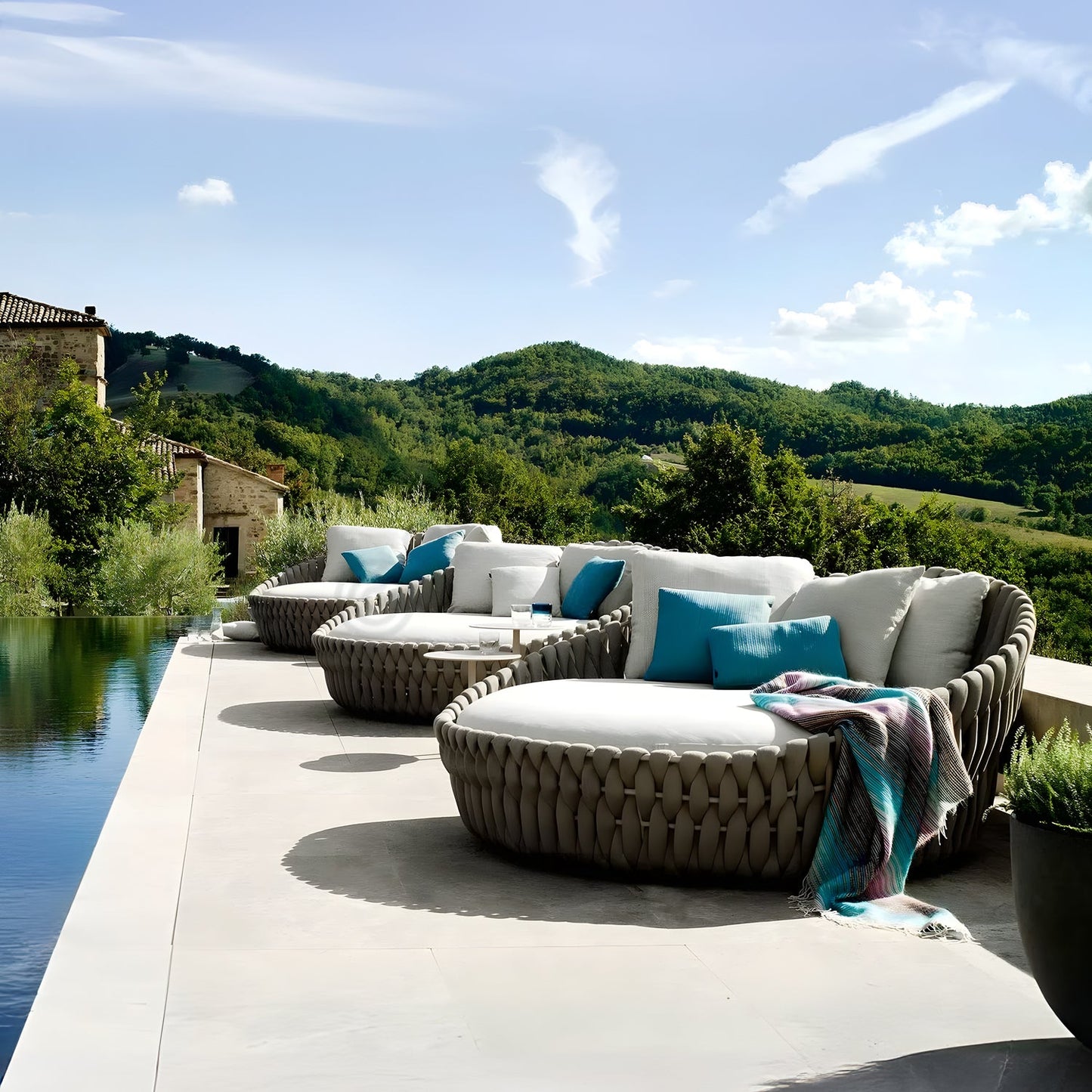 Experience an outdoor oasis with Landmark Decors Mediterranea Luxe Lounge featuring woven chairs, white cushions, and blue pillows by the infinity pool. A plaid blanket enhances coziness while scenic hills and a clear blue sky complete this luxurious retreat.