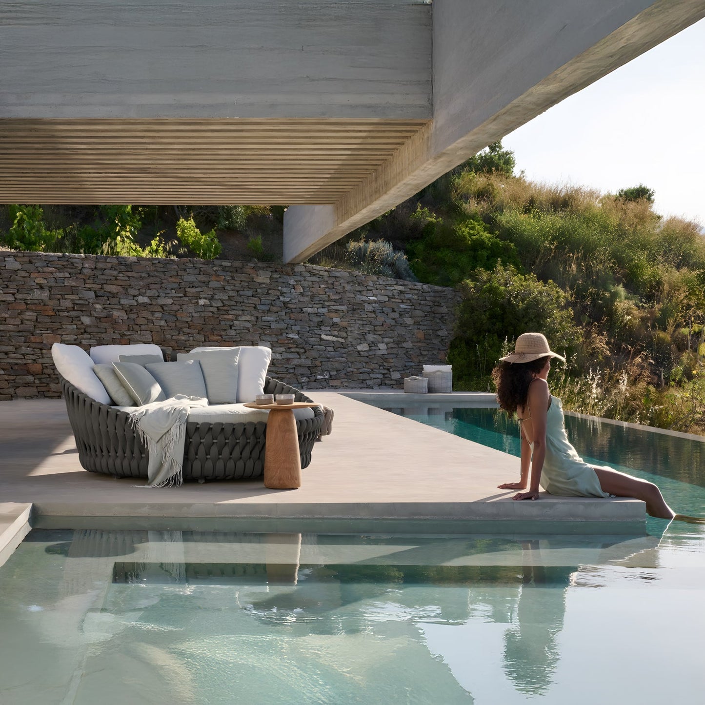 A woman in a sun hat relaxes by a pool, dipping her feet in the water next to the plush Mediterranea Luxe Lounge by Landmark Decor. Surrounded by stone walls and lush greenery beneath a modern concrete roof, this setting creates an inviting and luxurious outdoor retreat.