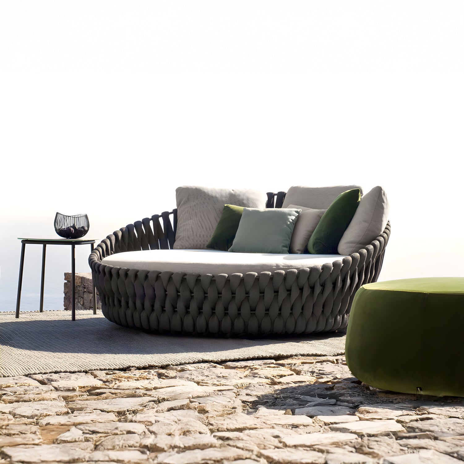 Nestled in a luxurious retreat, the outdoor oasis showcases Landmark Decors Mediterranea Luxe Lounge, a large round woven chair with green and white cushions. A nearby black side table on the stone patio holds a decorative glass piece, offering breathtaking sky views in this serene space.