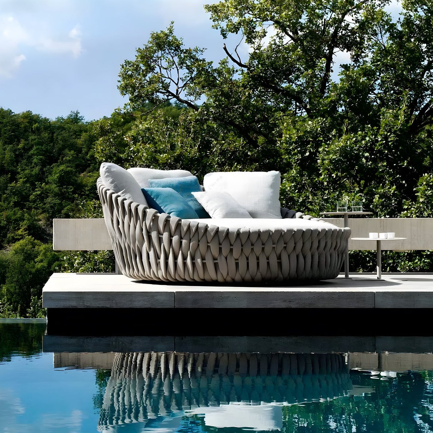The Mediterranea Luxe Lounge by Landmark Decor is a luxurious round daybed with woven design and white cushions, creating a perfect retreat on a sunlit stone patio by a tranquil pool, surrounded by lush trees and under a clear blue sky. A modern side table completes this idyllic outdoor space.