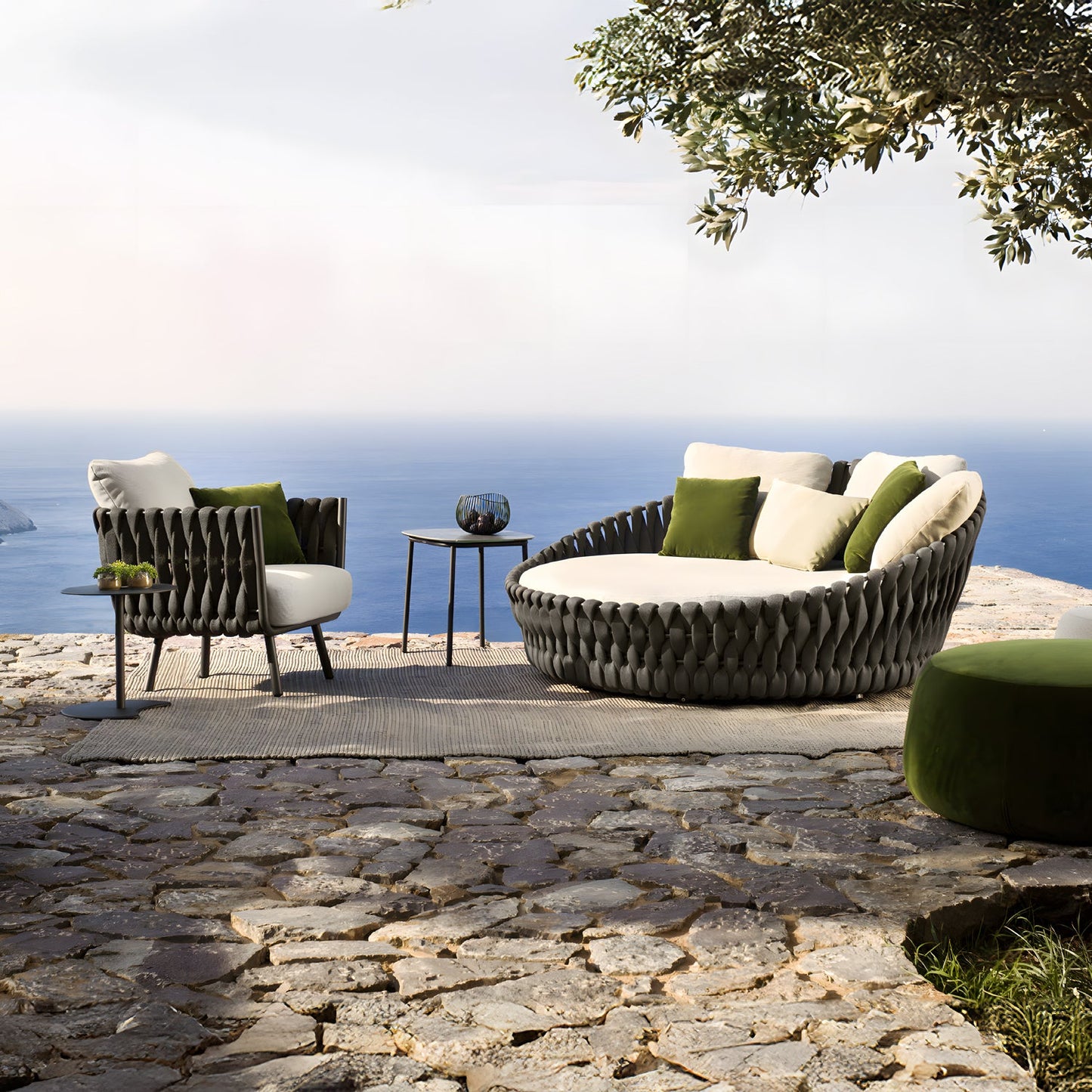 The outdoor patio is a luxurious retreat featuring the Mediterranea Luxe Lounge by Landmark Decor, with a woven furniture set including a large circular sofa and chair. Adorned with white and green cushions, it sits on a stone terrace overlooking the ocean, enhanced by a nearby olive tree.