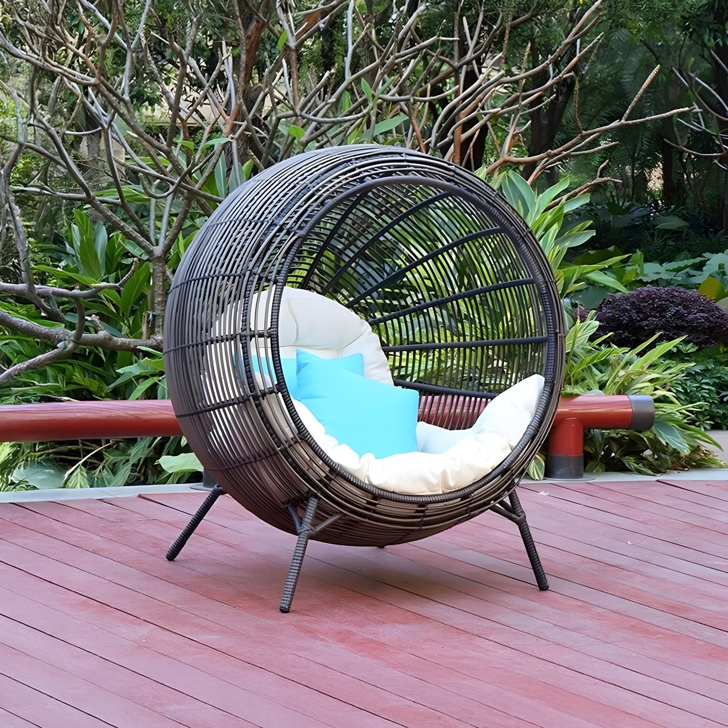 The Sphere Retreat Lounge by Landmark Decor, a modern wicker chair with white and blue cushions, graces the wooden deck of a lush garden. This inviting outdoor space, surrounded by verdant plants and trees, transforms into a luxurious retreat perfect for unwinding.