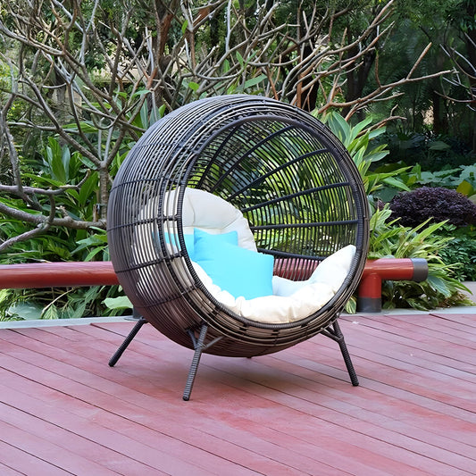 The Sphere Retreat Lounge by Landmark Decor, a modern wicker chair with white and blue cushions, graces the wooden deck of a lush garden. This inviting outdoor space, surrounded by verdant plants and trees, transforms into a luxurious retreat perfect for unwinding.