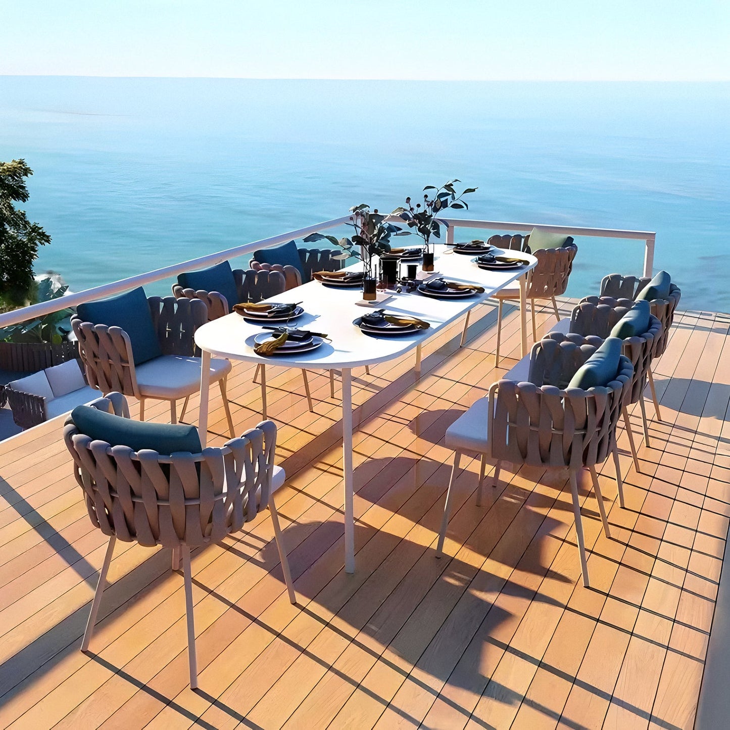 Indulge in alfresco dining on a wooden patio with Landmark Decors Coastal Woven Collection. A long white table, artfully set with plates, glasses, and cutlery, is surrounded by weather-resistant braided chairs. The clear blue sky and serene waters enhance the tranquil atmosphere.