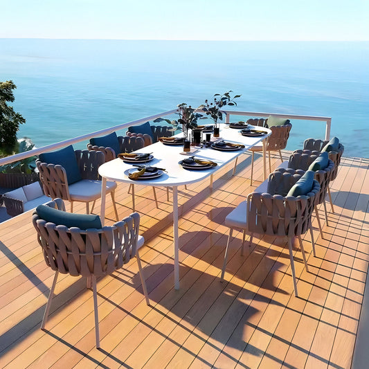 Indulge in alfresco dining on a wooden patio with Landmark Decors Coastal Woven Collection. A long white table, artfully set with plates, glasses, and cutlery, is surrounded by weather-resistant braided chairs. The clear blue sky and serene waters enhance the tranquil atmosphere.