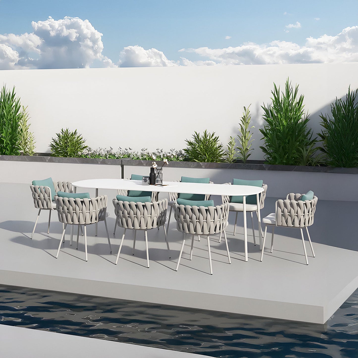 A modern alfresco dining area showcases the Coastal Woven Collection by Landmark Decor, with a long white table and woven chairs with blue cushions. The weather-resistant platform overlooks a narrow pool, bordered by green plants against a white wall beneath a partly cloudy sky.