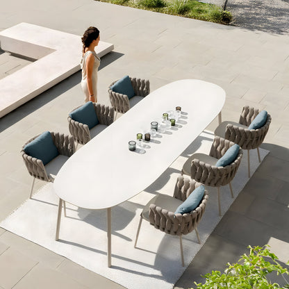 A person in a light dress walks by the Coastal Woven Collection table from Landmark Decor, set for alfresco dining with candles and glasses. Surrounding the weather-resistant table are eight gray chairs with teal cushions on a white rug outdoors on a stone patio.