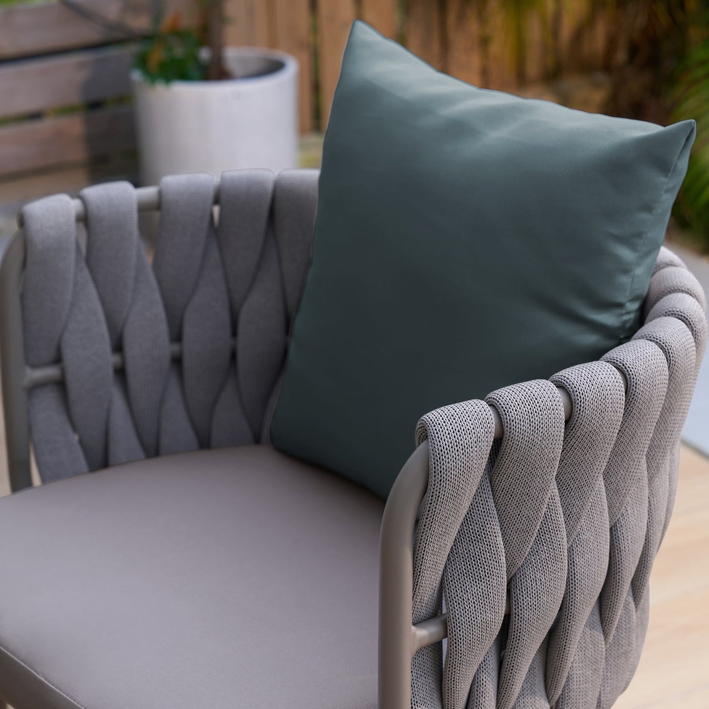 The Landmark Decor Coastal Woven Collection features a stylish, weather-resistant outdoor chair with a woven grey fabric design and sleek metal frame, topped with a large dark green cushion. In the background are a wooden bench and white pot plant, ideal for alfresco dining settings.