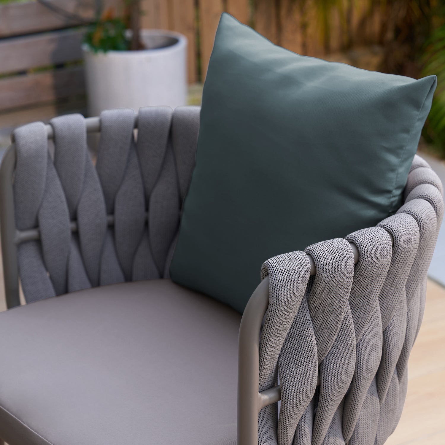 The Landmark Decor Coastal Woven Collection features a stylish, weather-resistant outdoor chair with a woven grey fabric design and sleek metal frame, topped with a large dark green cushion. In the background are a wooden bench and white pot plant, ideal for alfresco dining settings.