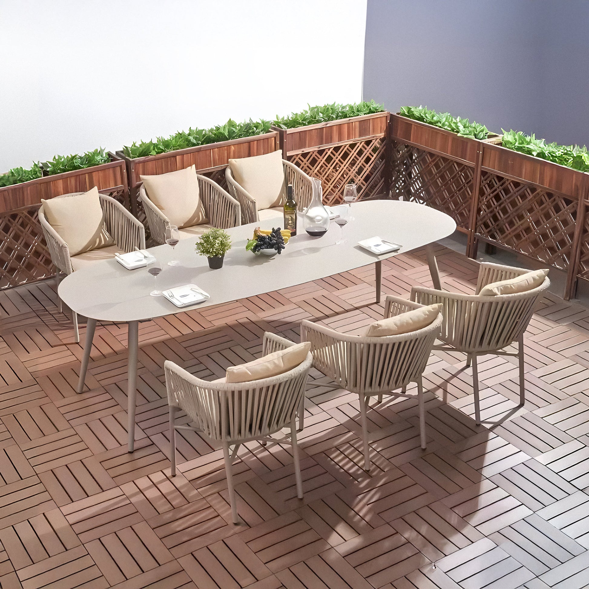Alfresco dining setup on a wooden patio featuring Landmark Decors St. Tropez Collection: a modern white, weather-resistant table with eight rope-woven chairs. The table is decorated with a plant, wine bottle, glasses, and fruit, while wooden planters with green plants line the background.