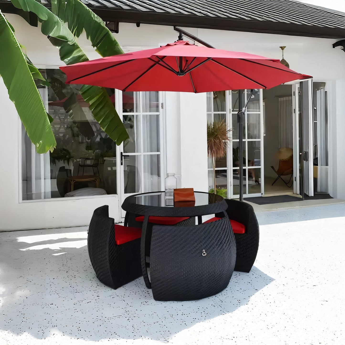 The Midnight Riviera Collection by Landmark Decor features a round glass table with four black wicker chairs and red cushions, all shaded by a large red umbrella. Ideal for alfresco dining, it sits on a white stone patio surrounded by banana plants and overlooks the houses open glass doors.