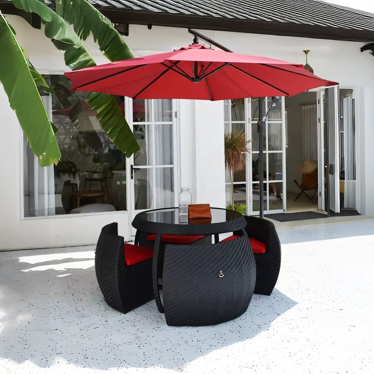 The Midnight Riviera Collection by Landmark Decor features a round glass table with four black wicker chairs and red cushions, all shaded by a large red umbrella. Ideal for alfresco dining, it sits on a white stone patio surrounded by banana plants and overlooks the houses open glass doors.