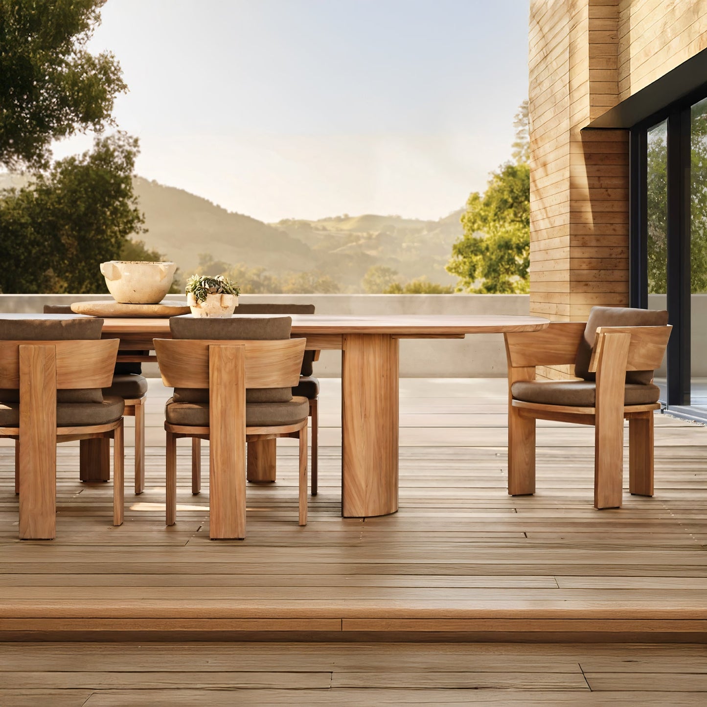 The Timber Serenity Collection by Landmark Decor offers a spacious outdoor patio setup for alfresco dining with a long wooden table and matching chairs. Surrounded by lush greenery and scenic hills, it’s connected to a modern building with large glass windows, ideal for sunny days.