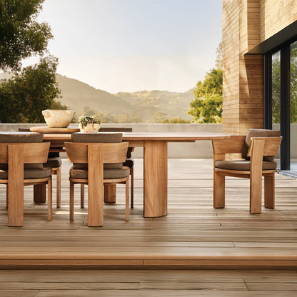 The Timber Serenity Collection by Landmark Decor offers a spacious outdoor patio setup for alfresco dining with a long wooden table and matching chairs. Surrounded by lush greenery and scenic hills, it’s connected to a modern building with large glass windows, ideal for sunny days.