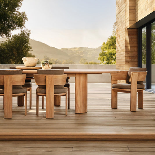 The Timber Serenity Collection by Landmark Decor offers a spacious outdoor patio setup for alfresco dining with a long wooden table and matching chairs. Surrounded by lush greenery and scenic hills, it’s connected to a modern building with large glass windows, ideal for sunny days.