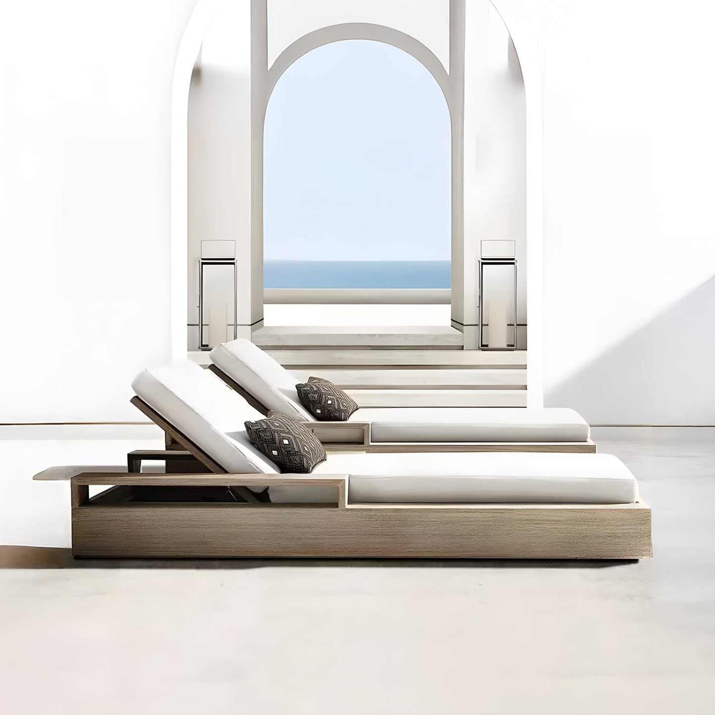 Two Landmark Decor Santorini Serenity Loungers with off-white cushions and patterned pillows sit in a bright, minimalist space. An archway reveals a clear blue sky and ocean view. Steps lead to the outdoor living area, where soft shadows enhance the serene setting crafted from premium materials.