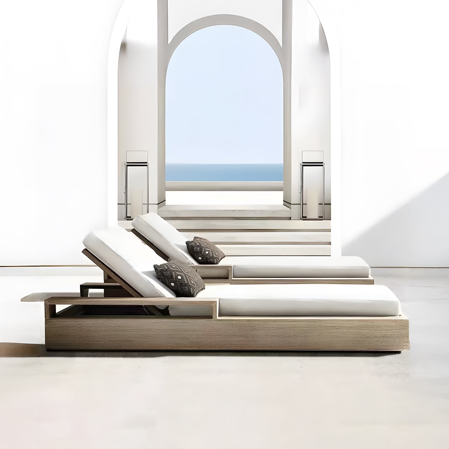 Two Landmark Decor Santorini Serenity Loungers with off-white cushions and patterned pillows sit in a bright, minimalist space. An archway reveals a clear blue sky and ocean view. Steps lead to the outdoor living area, where soft shadows enhance the serene setting crafted from premium materials.