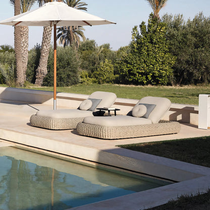 Two Marbella Retreat Loungers by Landmark Decor are arranged poolside with cushions. A large umbrella provides shade, complementing the luxurious atmosphere. Palm trees and greenery add to the serene outdoor setting, perfect for relaxing outdoor living.