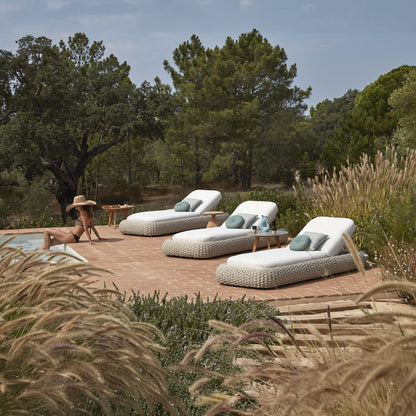 Amid lush greenery, a scenic outdoor living area boasts three Marbella Retreat Loungers by Landmark Decor on a brick patio. Someone in a large sun hat relaxes on one lounger, while cushions and small tables with decor complete this luxurious retreat.