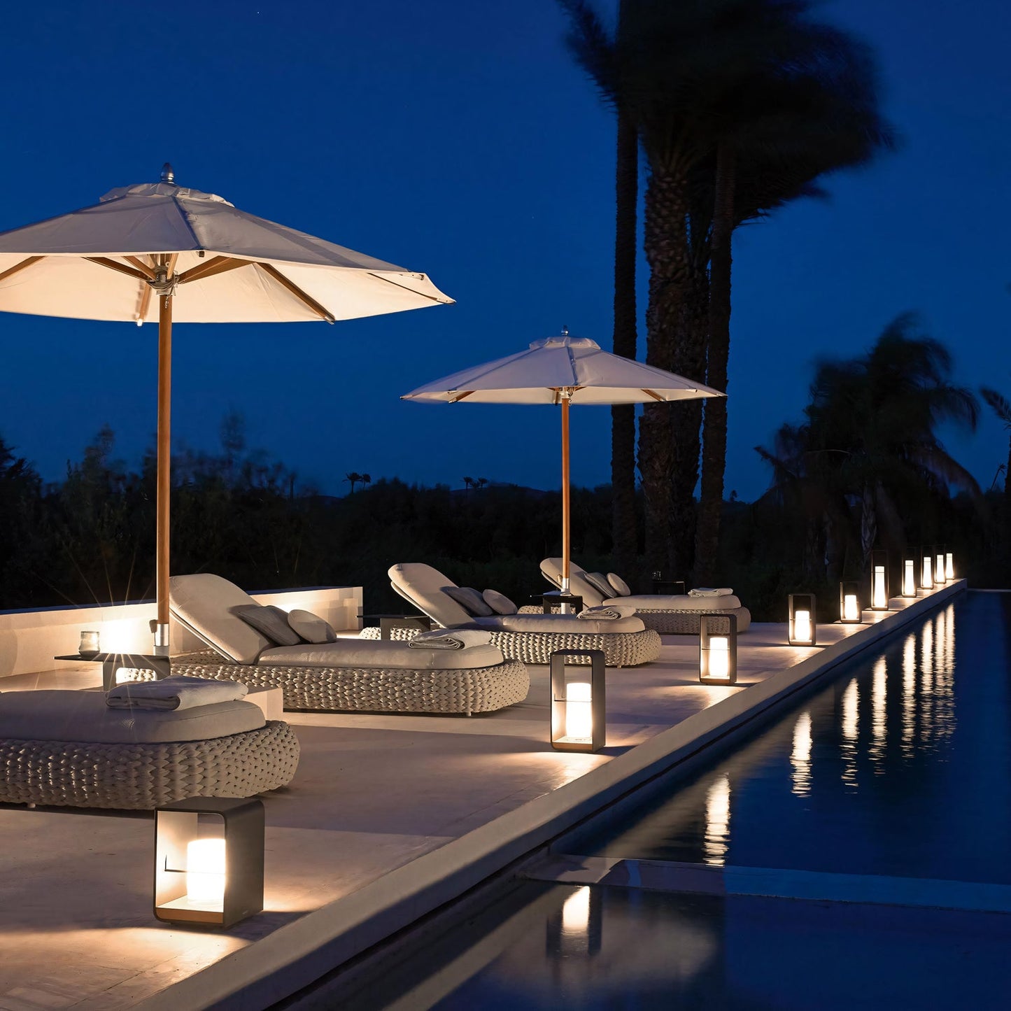 In this luxurious retreat, the poolside at night features Marbella Retreat Loungers by Landmark Decor under large umbrellas. Lanterns provide a warm glow beside the pool, and palm trees are beautifully silhouetted against the deep blue sky, creating an ideal outdoor atmosphere.