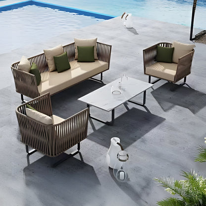 The outdoor lounge boasts the Sierra Luxe Collection by Landmark Decor with a wicker sofa, two armchairs with beige and green weather-resistant cushions, a white rectangular table, and two decorative vases, all overlooking a shimmering blue pool—ideal for relaxation in style.