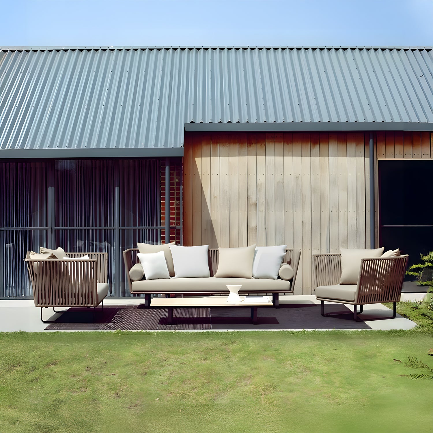 On a wooden deck, the Sierra Luxe Collection by Landmark Decor features a modern outdoor sofa set with weather-resistant cushions, including light-colored sofas and armchairs. A building with a metal roof and wooden walls complements the scene, surrounded by lush greenery.