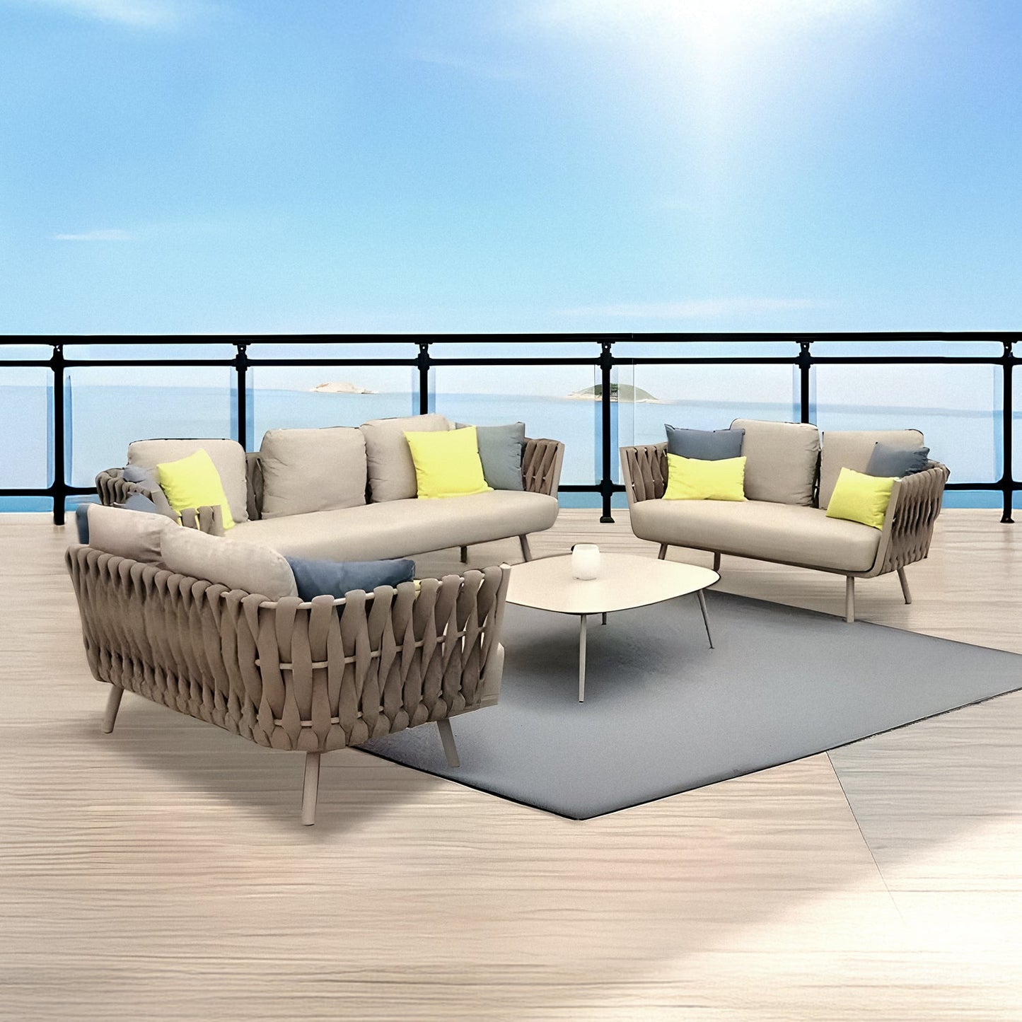 The outdoor terrace showcases the Landmark Decor Elysian Breeze Collection, featuring two beige sofas with weather-resistant yellow and green cushions, a triangular coffee table on a gray rug. A railing offers a view of the calm blue sea under a clear sky.