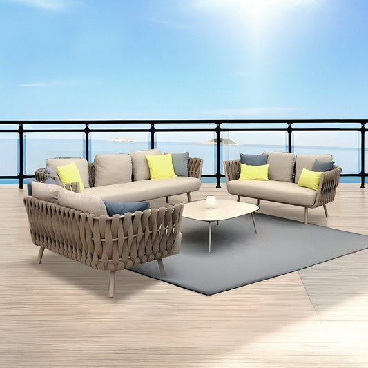 The outdoor terrace showcases the Landmark Decor Elysian Breeze Collection, featuring two beige sofas with weather-resistant yellow and green cushions, a triangular coffee table on a gray rug. A railing offers a view of the calm blue sea under a clear sky.