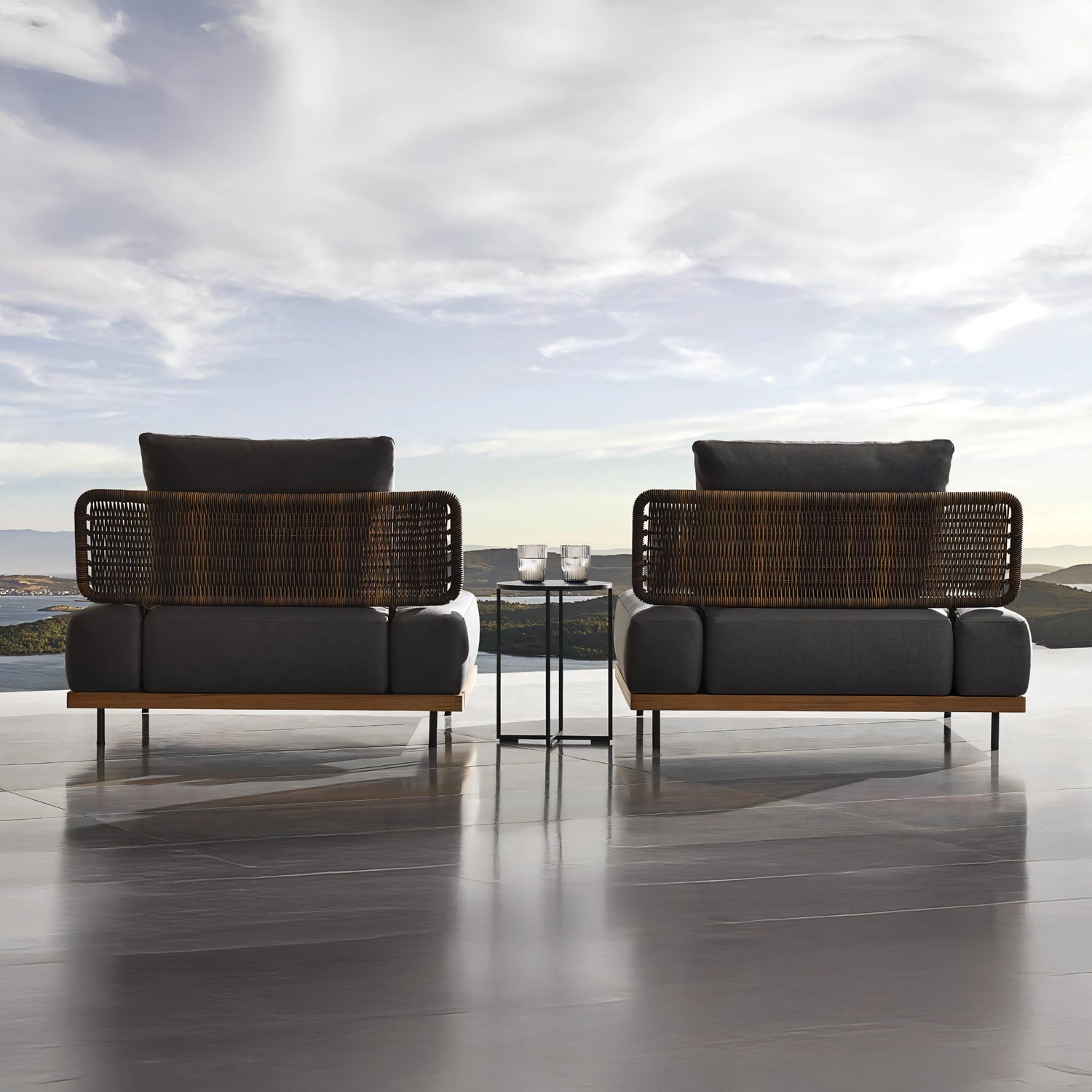 Two wicker and cushioned lounge chairs from the Oceanview Oasis Collection by Landmark Decor feature weather-resistant cushions, facing a mountain view and cloudy sky. A small table with candles completes the setting on the glossy patio floor, creating a perfect space for relaxation.