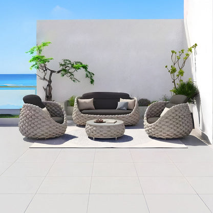 Outdoor patio featuring Landmark Decors Nordic Lux Collection: a woven outdoor sofa, two armchairs, and a round coffee table on a light rug. Potted plants adorn a white wall with an ocean view under the clear blue sky. Enjoy all-weather frames and weather-resistant cushions.