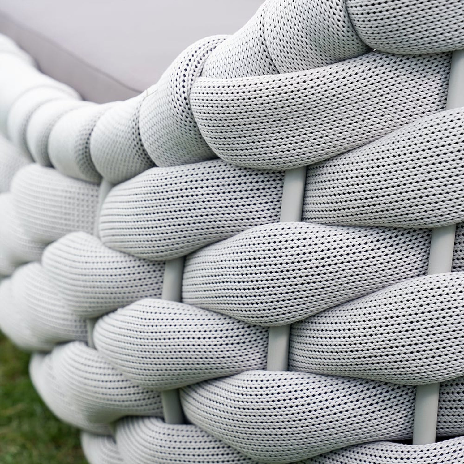 A close-up of the Nordic Lux Collection chair by Landmark Decor showcases its textured woven design with thick, light gray interlaced strands in a braided pattern. Its soft yet durable surface is enhanced by weather-resistant cushions, set against a blurred grassy background.