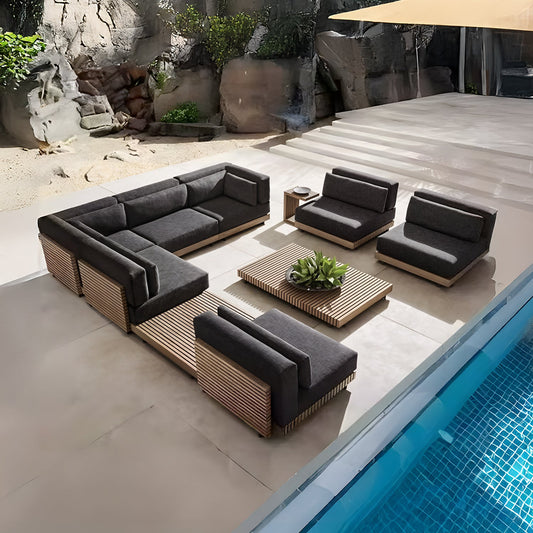 The outdoor seating area features the Landmark Decor Palazzo Tranquillo Collection, a modern modular sofa set with dark weather-resistant cushions and all-weather wooden frames. A square coffee table with a plant centerpiece completes the setting by the pool against rocky and lush green scenery.