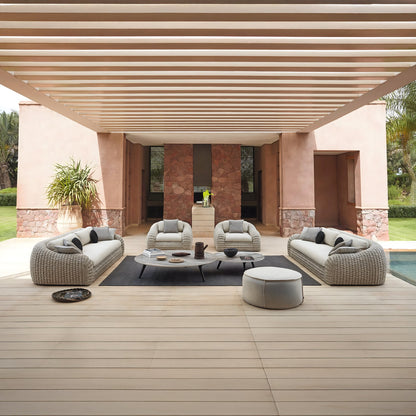 The outdoor patio showcases the Marbella Retreat Collection by Landmark Decor, featuring modern wicker sofas with weather-resistant cushions and a round ottoman on a wooden deck. A large rug and low table hold decorative items, beneath a beige pergola surrounded by potted plants, stone walls, and lush greenery.