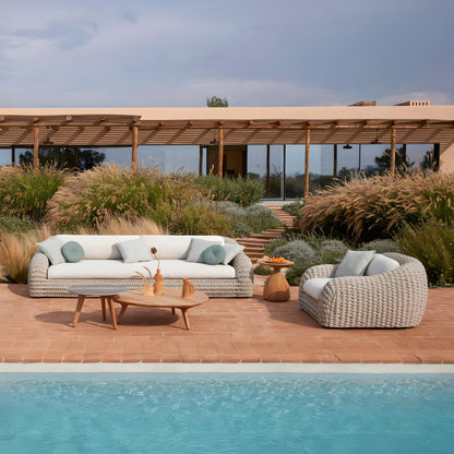 An outdoor patio showcasing Landmark Decors Marbella Retreat Collection, with woven sofas and armchairs featuring weather-resistant cushions, a wooden coffee table adorned with vases, and a side table by the pool. Surrounded by lush greenery is a modern house with expansive windows.