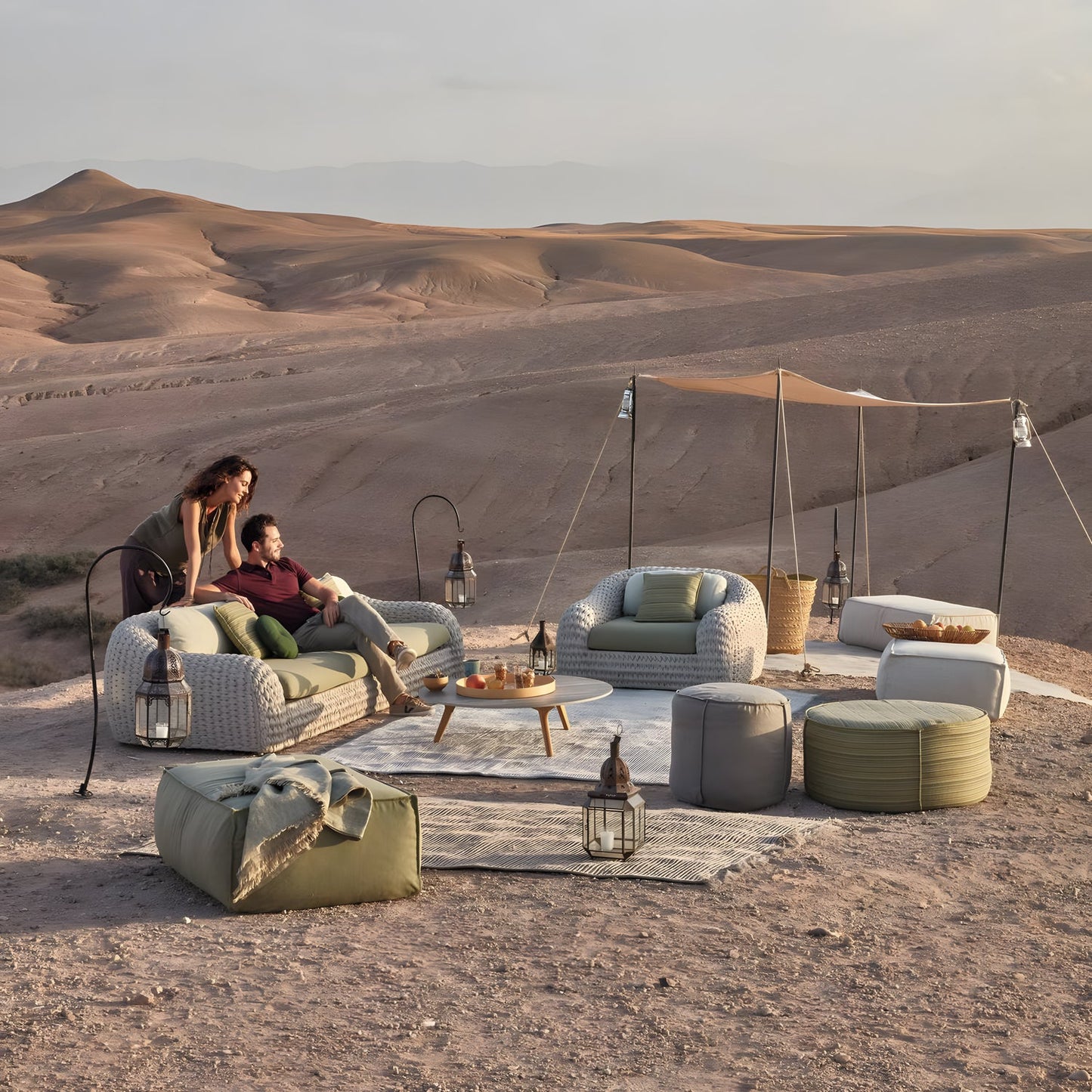 In a desert setting, a couple enjoys the Marbella Retreat Collection by Landmark Decor. Woven furniture with all-weather frames and weather-resistant cushions are complemented by lanterns and a canopy, against brown dunes under a cloudy sky.