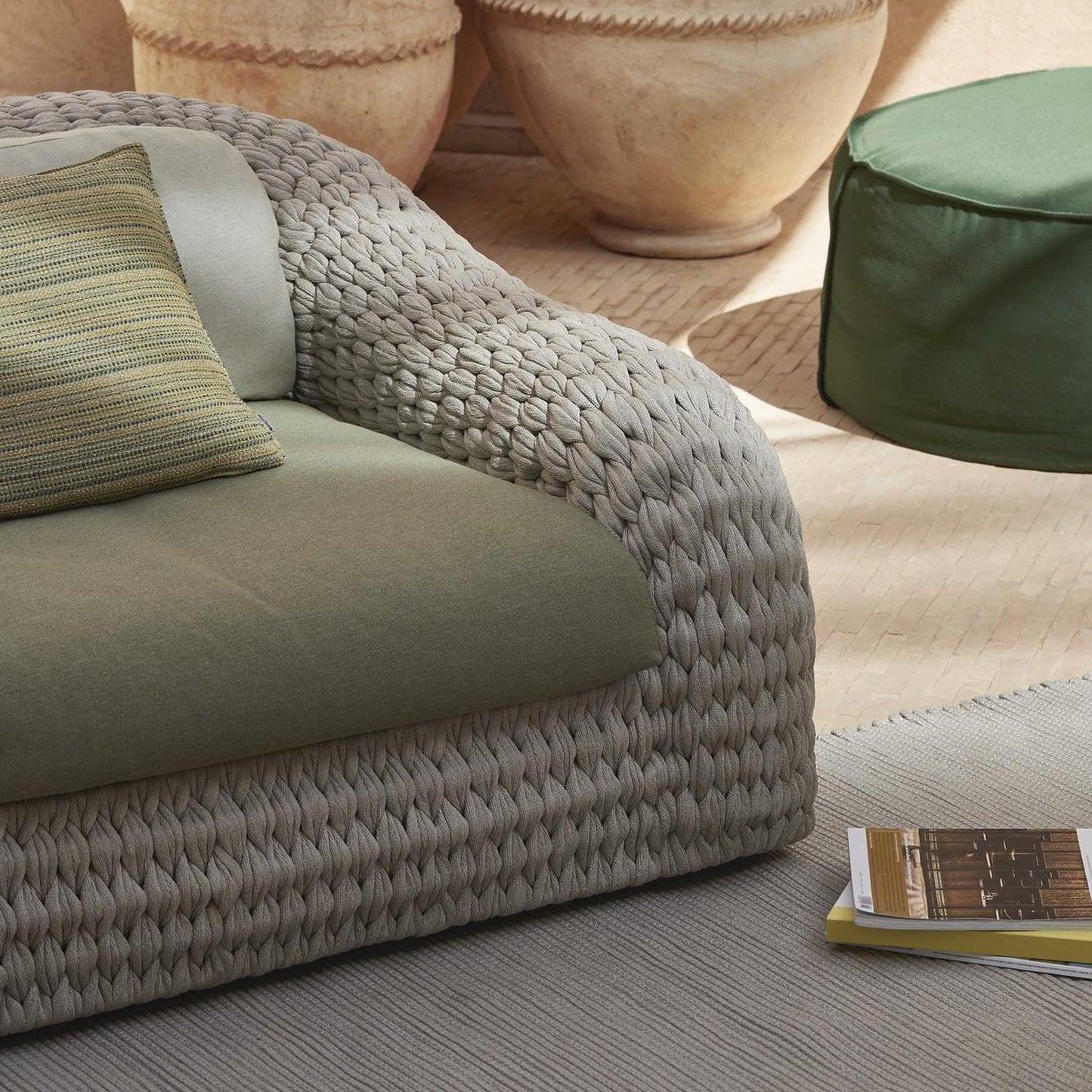 Create a cozy vibe with the Marbella Retreat Collection by Landmark Decor, featuring a gray woven outdoor sofa with weather-resistant green cushions, a round green ottoman, and a book on a gray rug. Large clay pots add an earthy touch for perfect breezy afternoons.