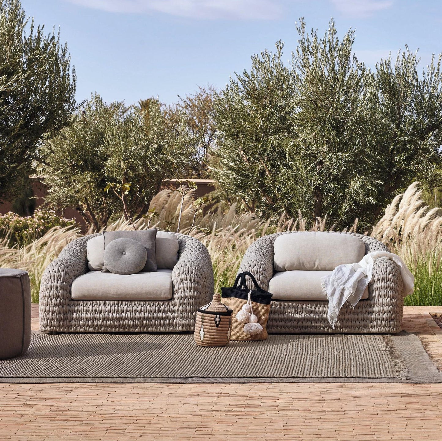 The Marbella Retreat Collection by Landmark Decor features two woven armchairs with light, weather-resistant cushions on a textured rug. A basket and black pom-pom tote sit between them, surrounded by tall grasses and trees against a blue sky, all framed in durable materials.