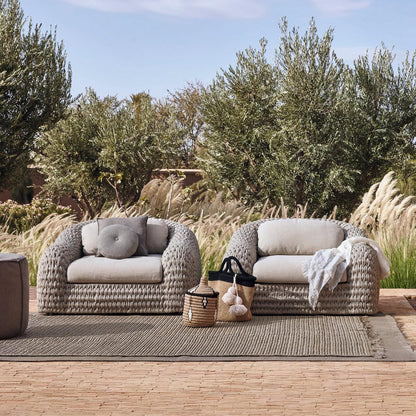The Marbella Retreat Collection by Landmark Decor features two woven armchairs with light, weather-resistant cushions on a textured rug. A basket and black pom-pom tote sit between them, surrounded by tall grasses and trees against a blue sky, all framed in durable materials.