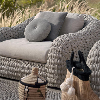The Marbella Retreat Collection by Landmark Decor features a close-up of an outdoor sofa with grey, weather-resistant cushions and a round pillow. Two woven baskets with lids sit on a textured rug, surrounded by grass and plants, emphasizing the all-weather frames’ lasting durability.