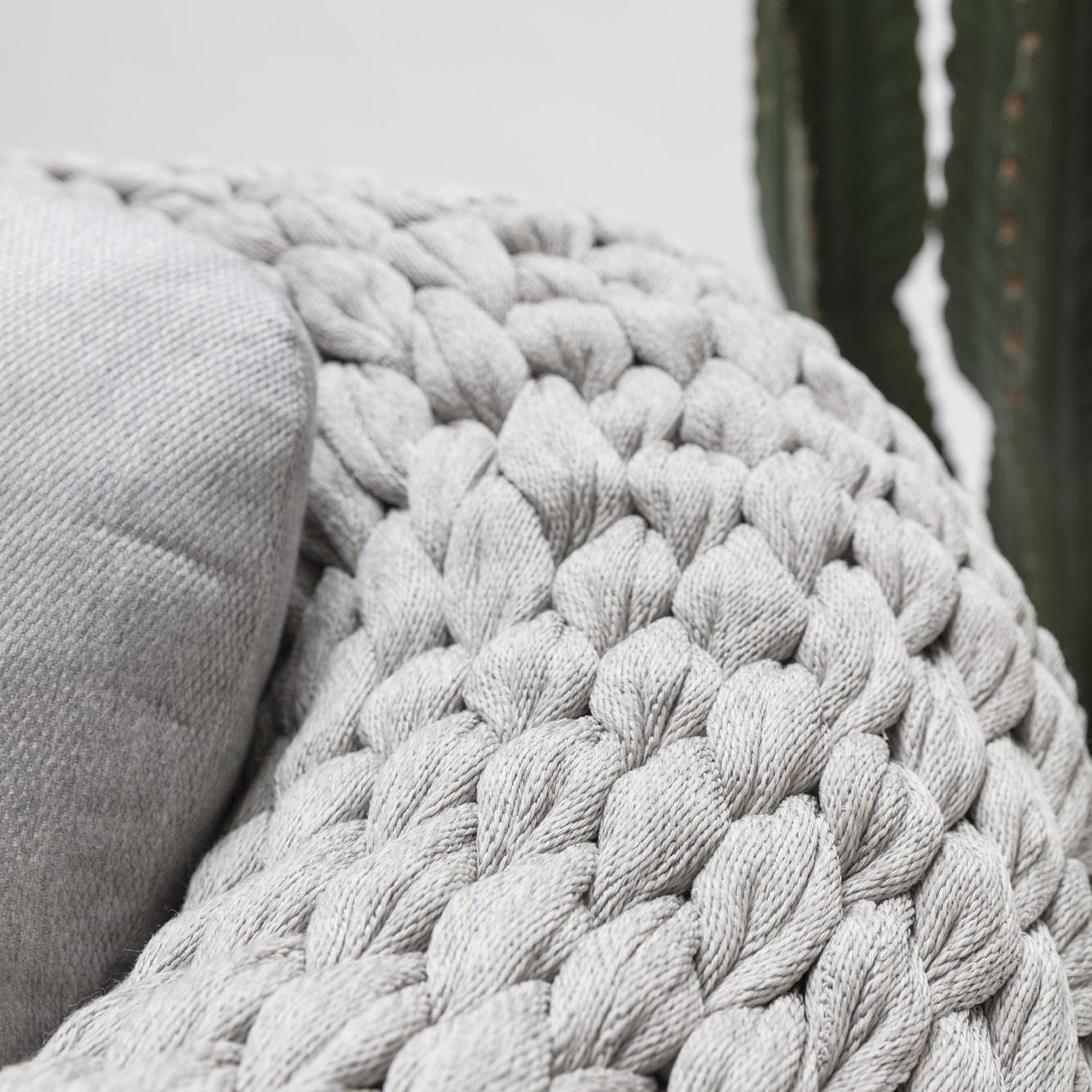 A gray chunky knit blanket from Landmark Decors Marbella Retreat Collection drapes over an outdoor sofa arm. A weather-resistant cushion peeks out beside it, while a tall green cactus contrasts with the soft textiles in the background.