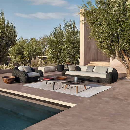 The Palermo Terra Collection by Landmark Decor features modern wicker sofas with weather-resistant cushions, two coffee tables, and a large rug, creating a stylish and comfortable poolside setting framed by olive trees and a beige building under the blue sky.